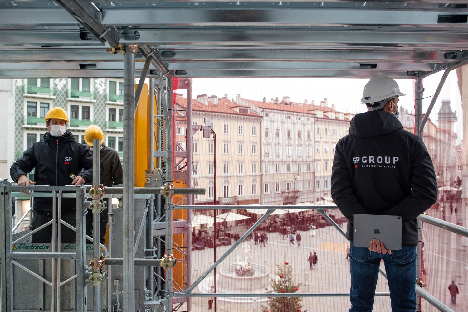 Two construction workers in hard hats survey a city square from a building scaffold during a renovation project.