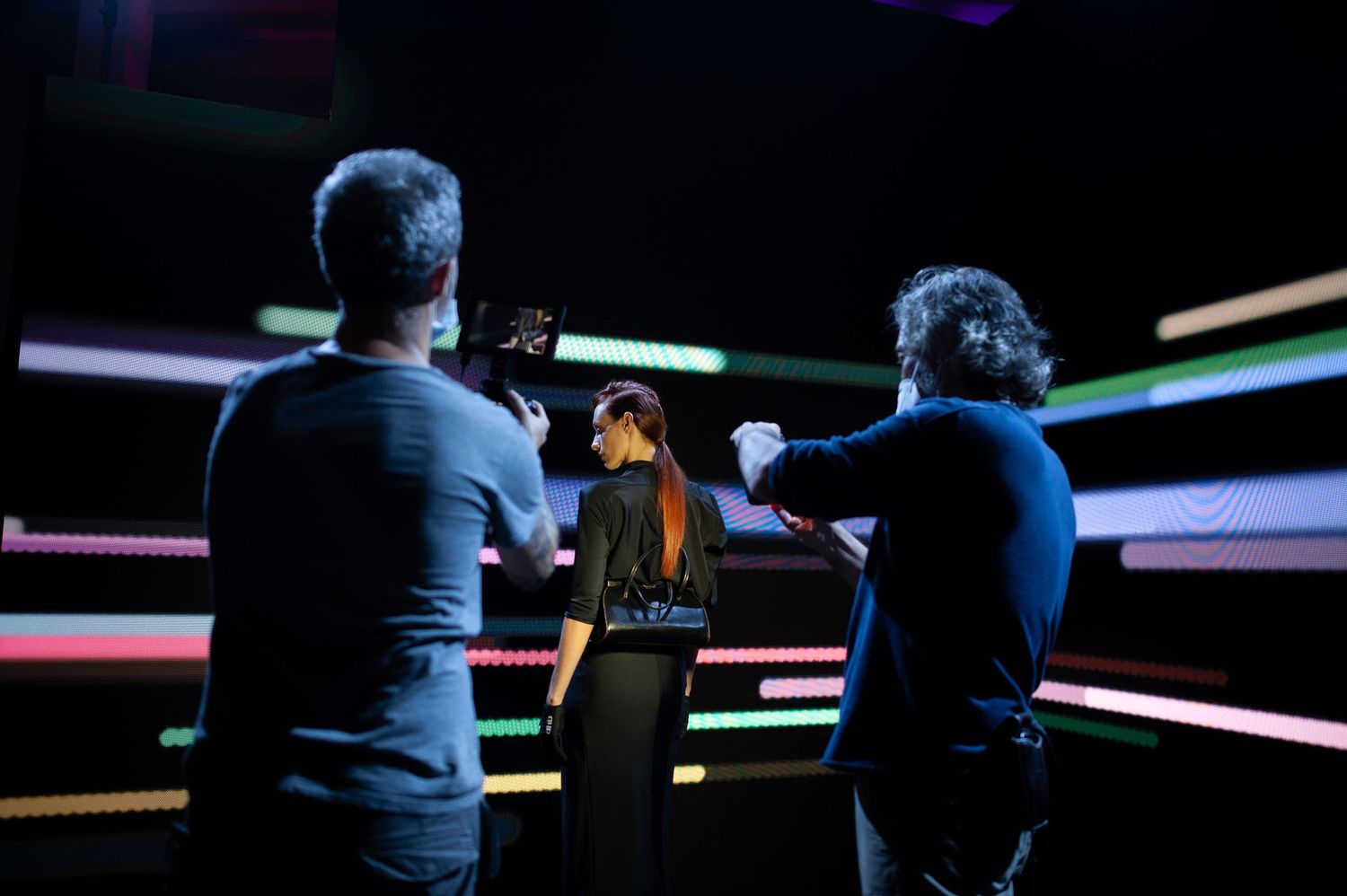 Three people stand in a dark room with colorful neon light strips, appearing to film or photograph a woman in black.