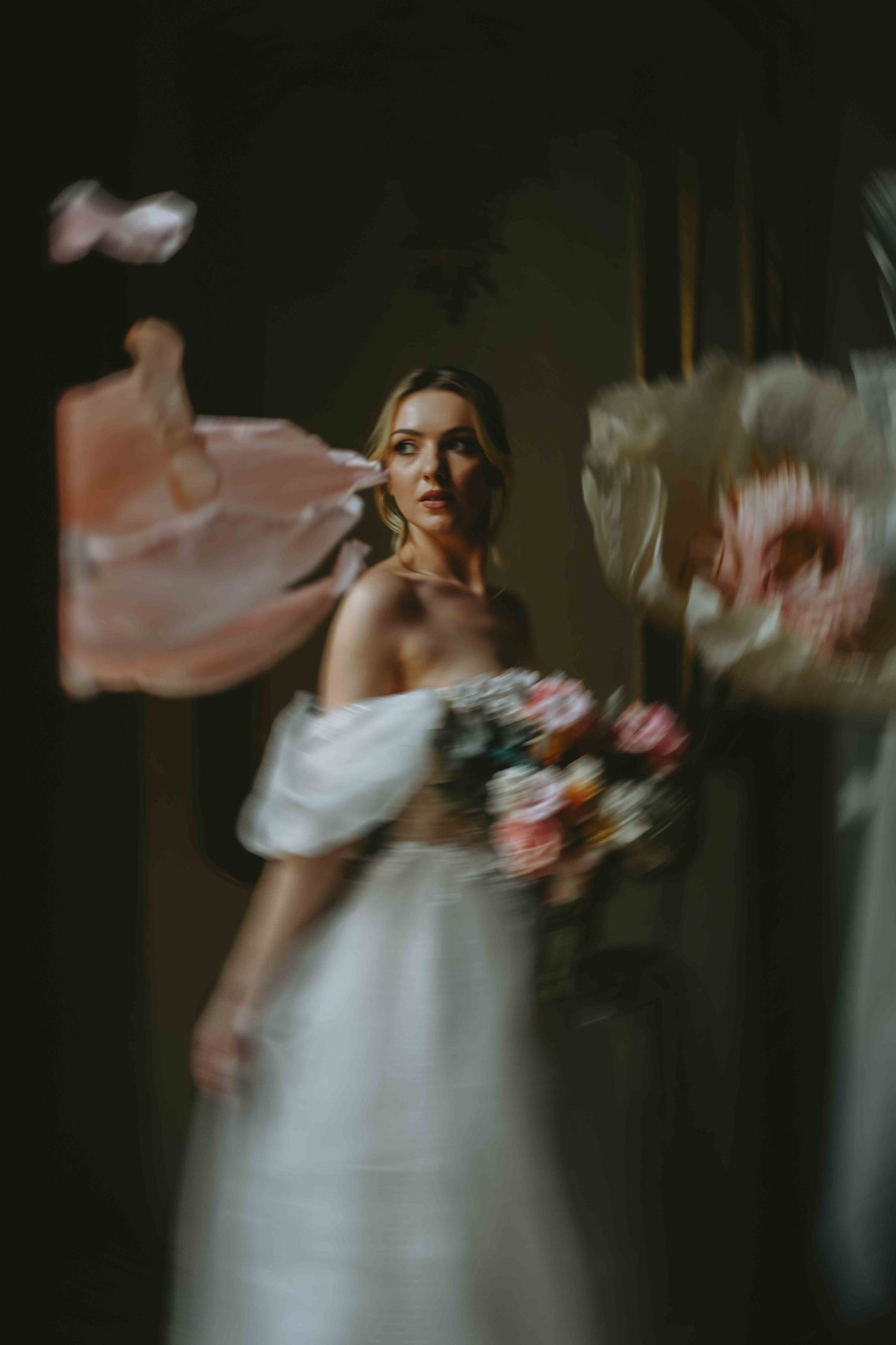 Bride in off-shoulder white gown holds floral bouquet, surrounded by blurred pink fabric in moody, artistic wedding photo.