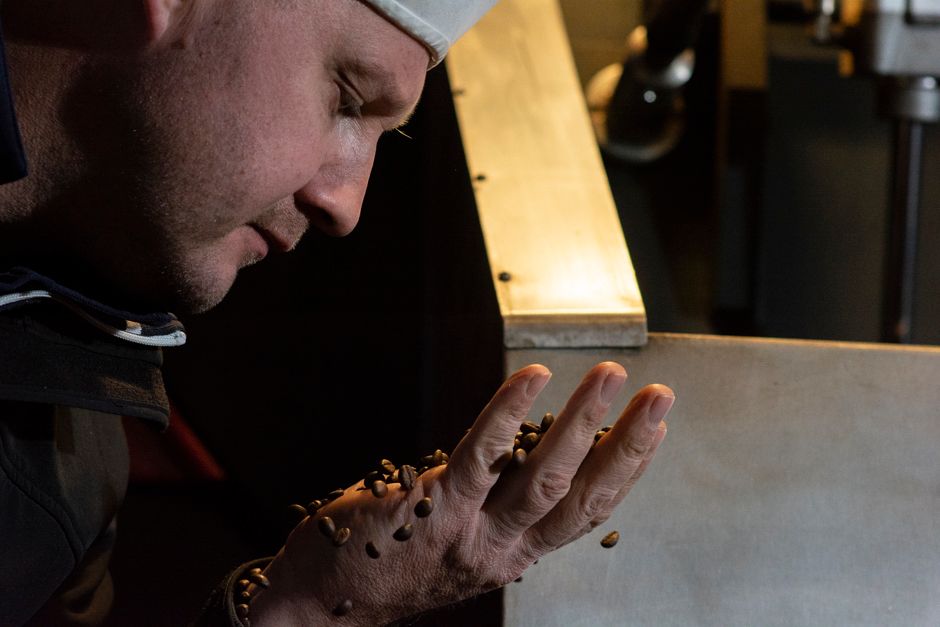 A man in a white cap closely examines roasted coffee beans held in his hands near industrial equipment.