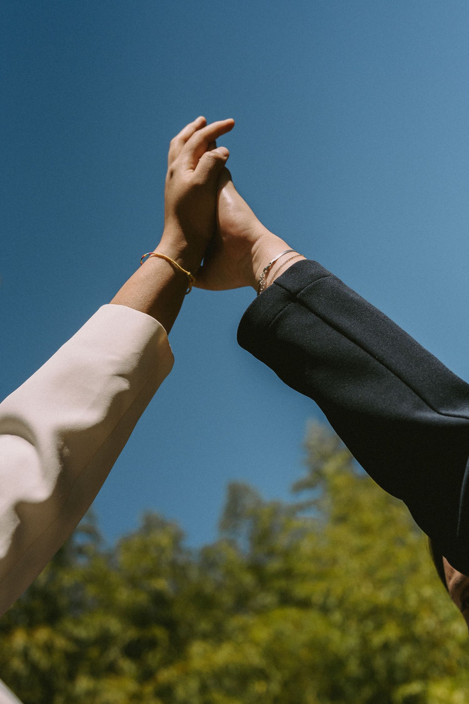 Two hands reaching up together against a blue sky, both wearing gold bracelets, symbolizing friendship and unity.