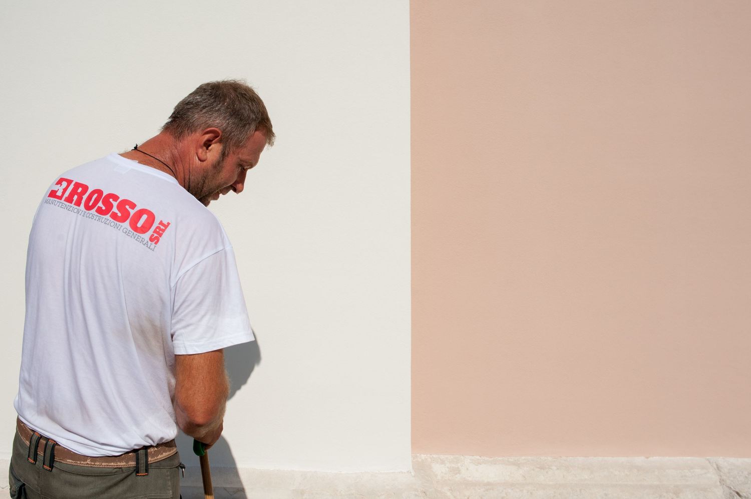 A man in a white Rosso branded t-shirt examines a freshly painted pink and white exterior wall.
