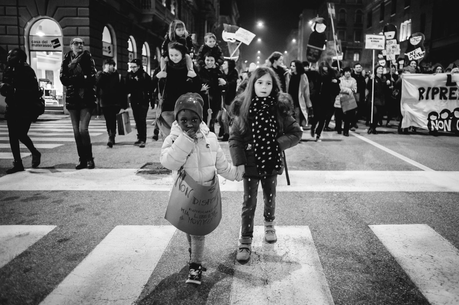 Two young girls hold hands at a crosswalk during a nighttime protest march in a black and white photo.