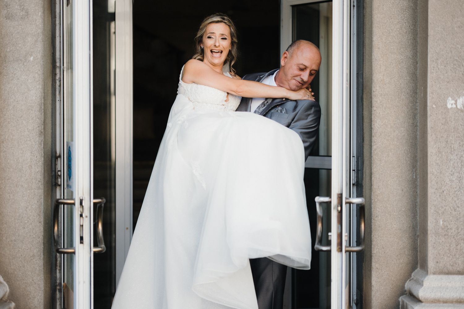 A bride in a white off-shoulder wedding gown stands in front of glass doors at an elegant venue.