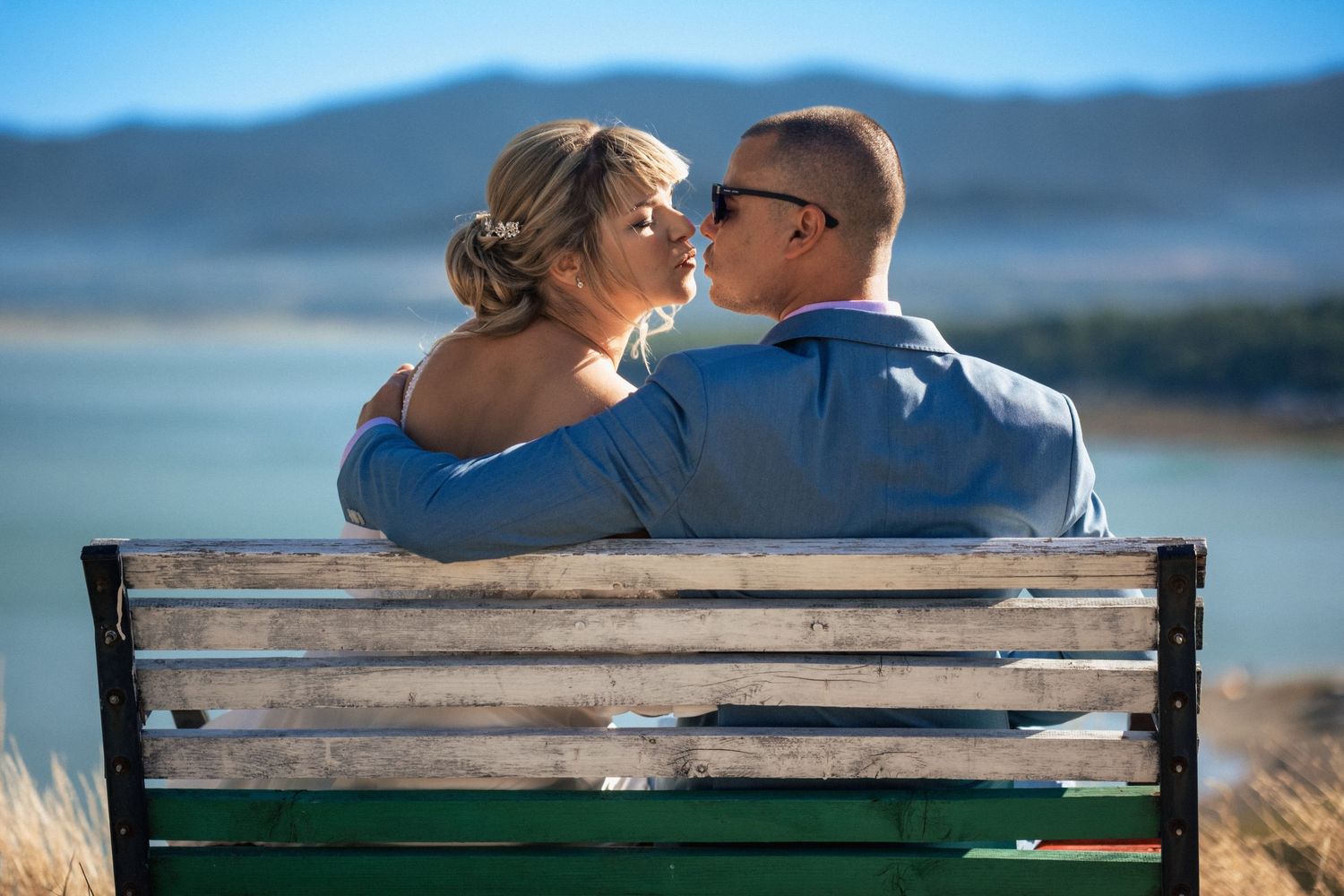 Couple sharing romantic moments on a wooden bench overlooking a scenic lake with mountains in the background.