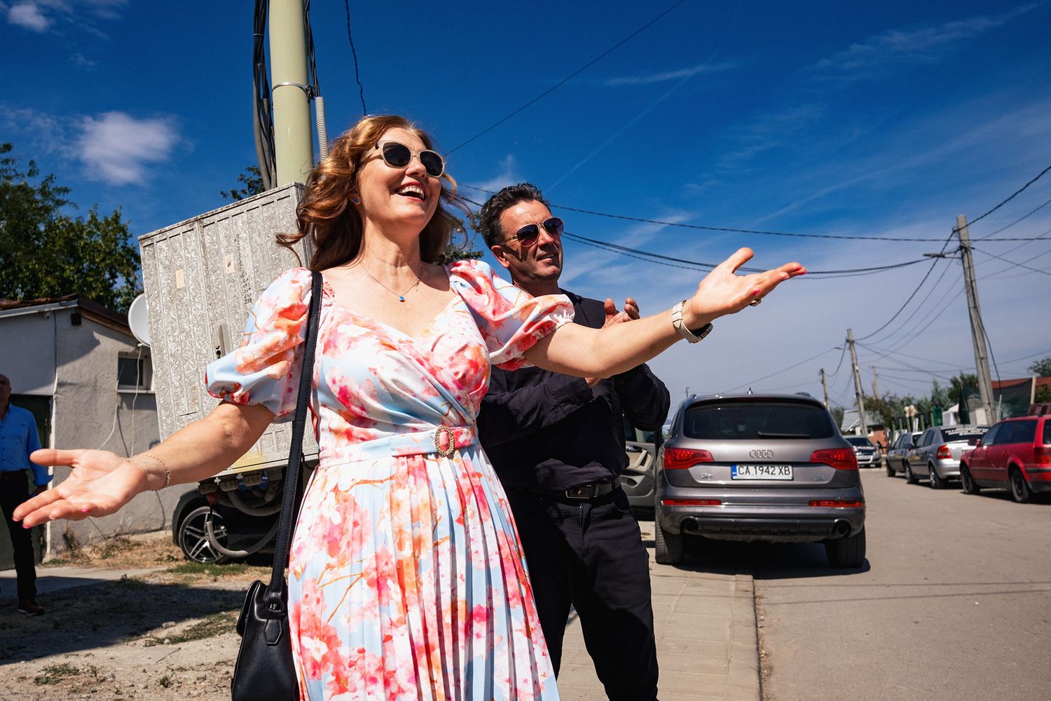 Wedding party dances and poses outdoors on a sunny day with cars in background.