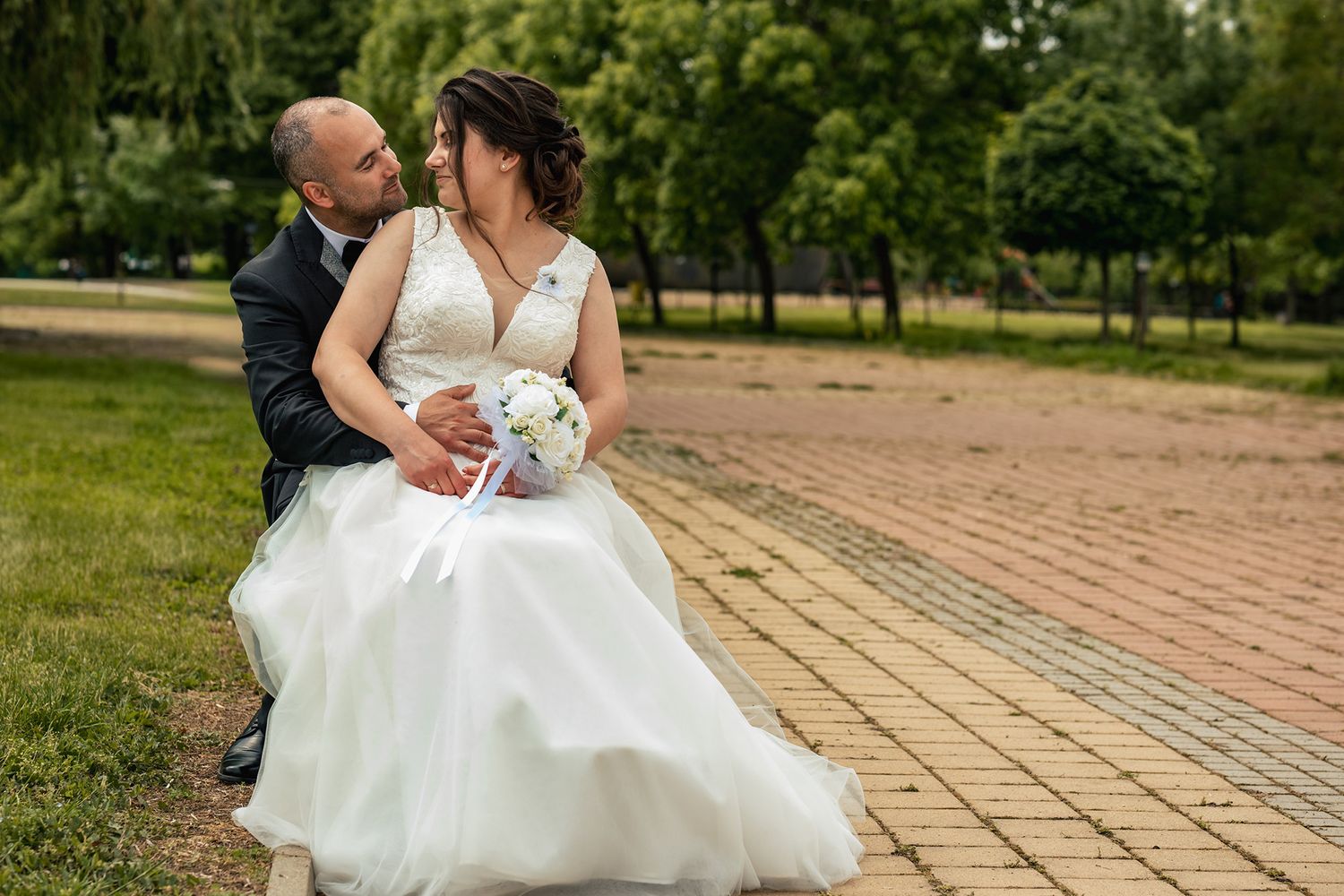 A couple in wedding attire sitting together on brick pavers in a park setting.