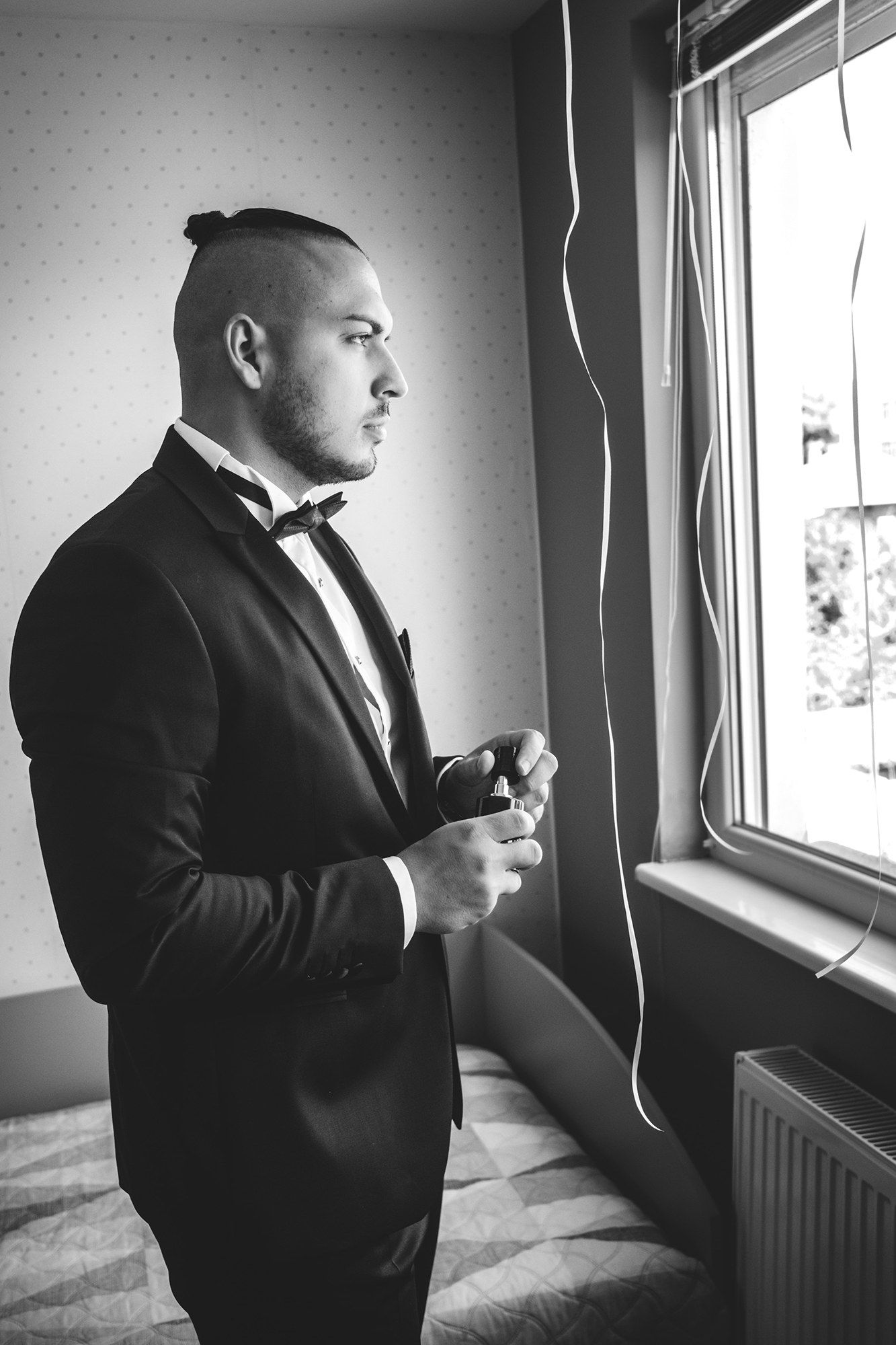 Black and white photos of groom getting ready by window in formal attire.