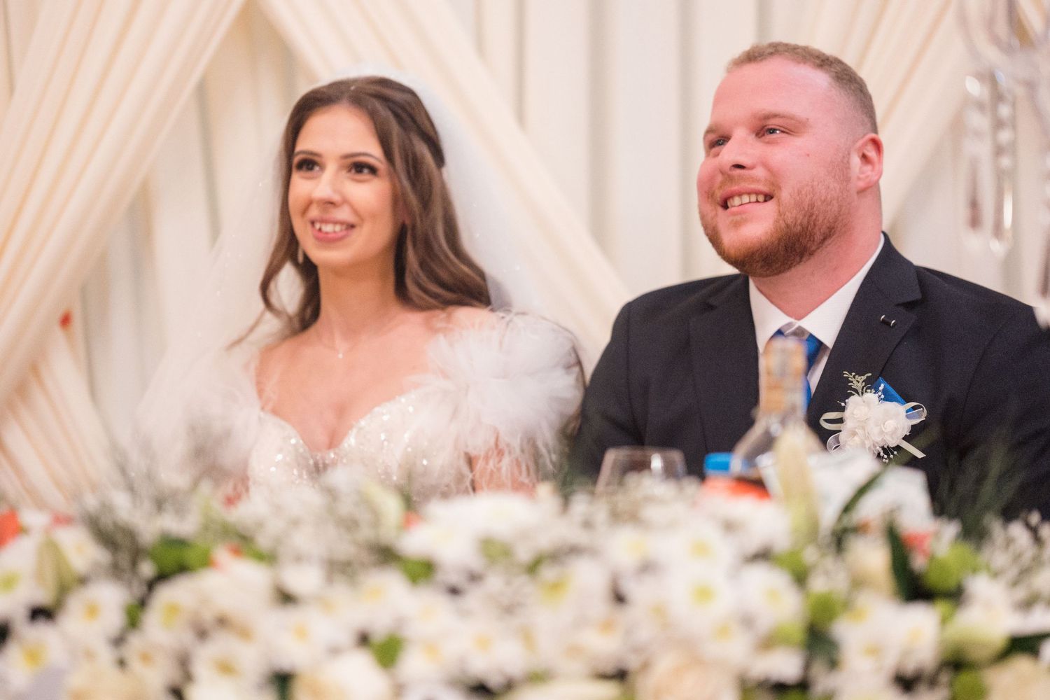 Wedding party members sit at a decorated head table with white floral centerpieces during a reception celebration.