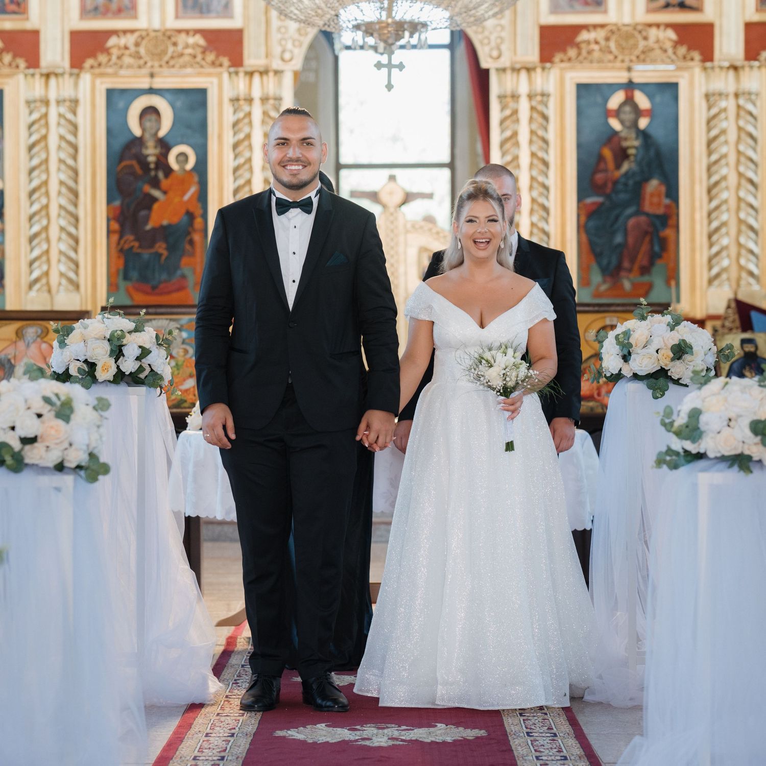 Orthodox church wedding ceremony with couple standing on ornate red carpet surrounded by religious icons and decorations.