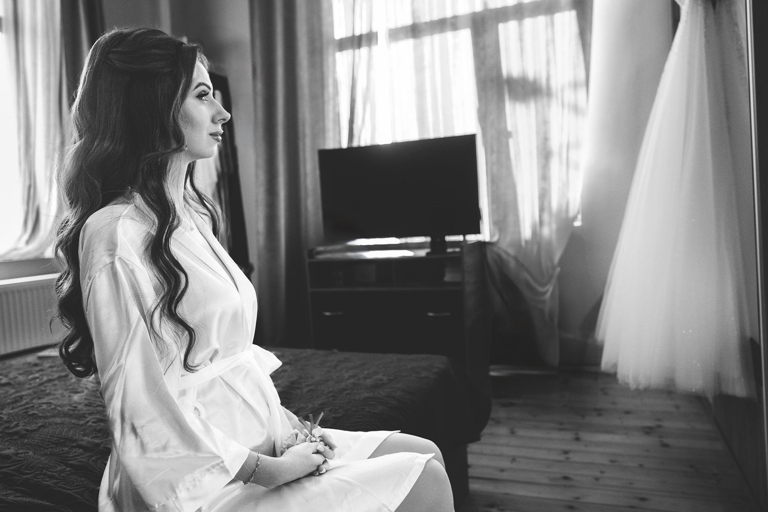 Bride in white robe sits contemplatively by window during wedding day preparations.