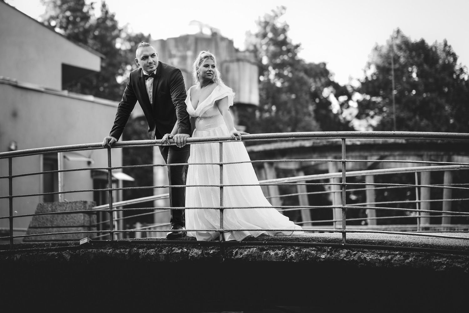 A black and white wedding photo of a couple standing together on a modern bridge railing.