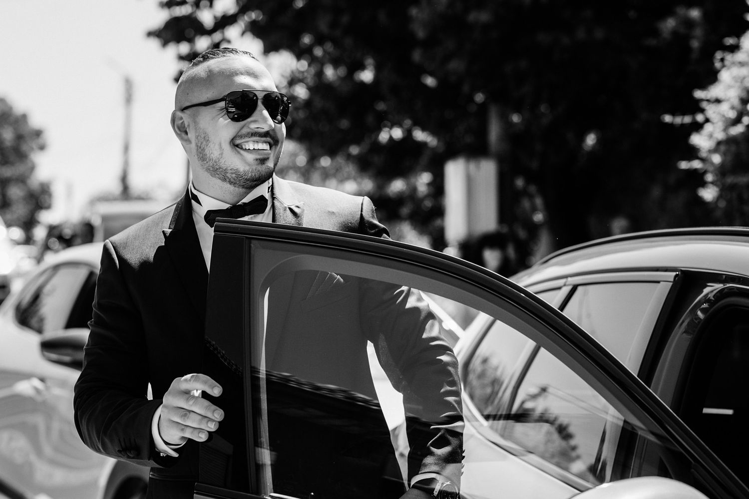 Groomsmen in sunglasses and suits smiling near a black car.