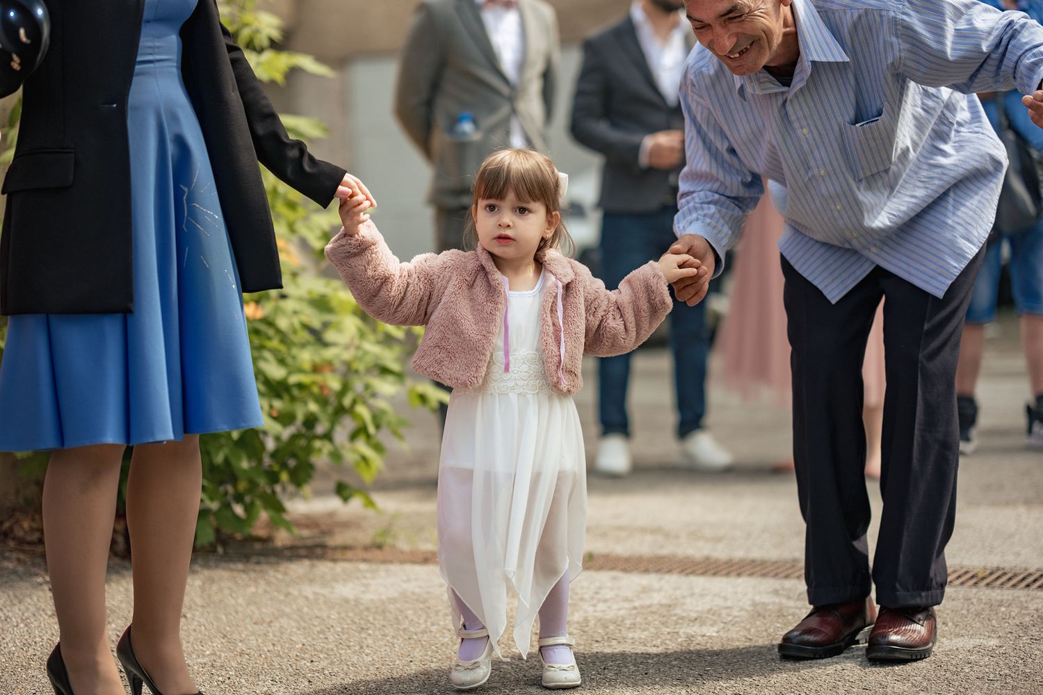 Small child in white outfit taking first steps with assistance during outdoor gathering.