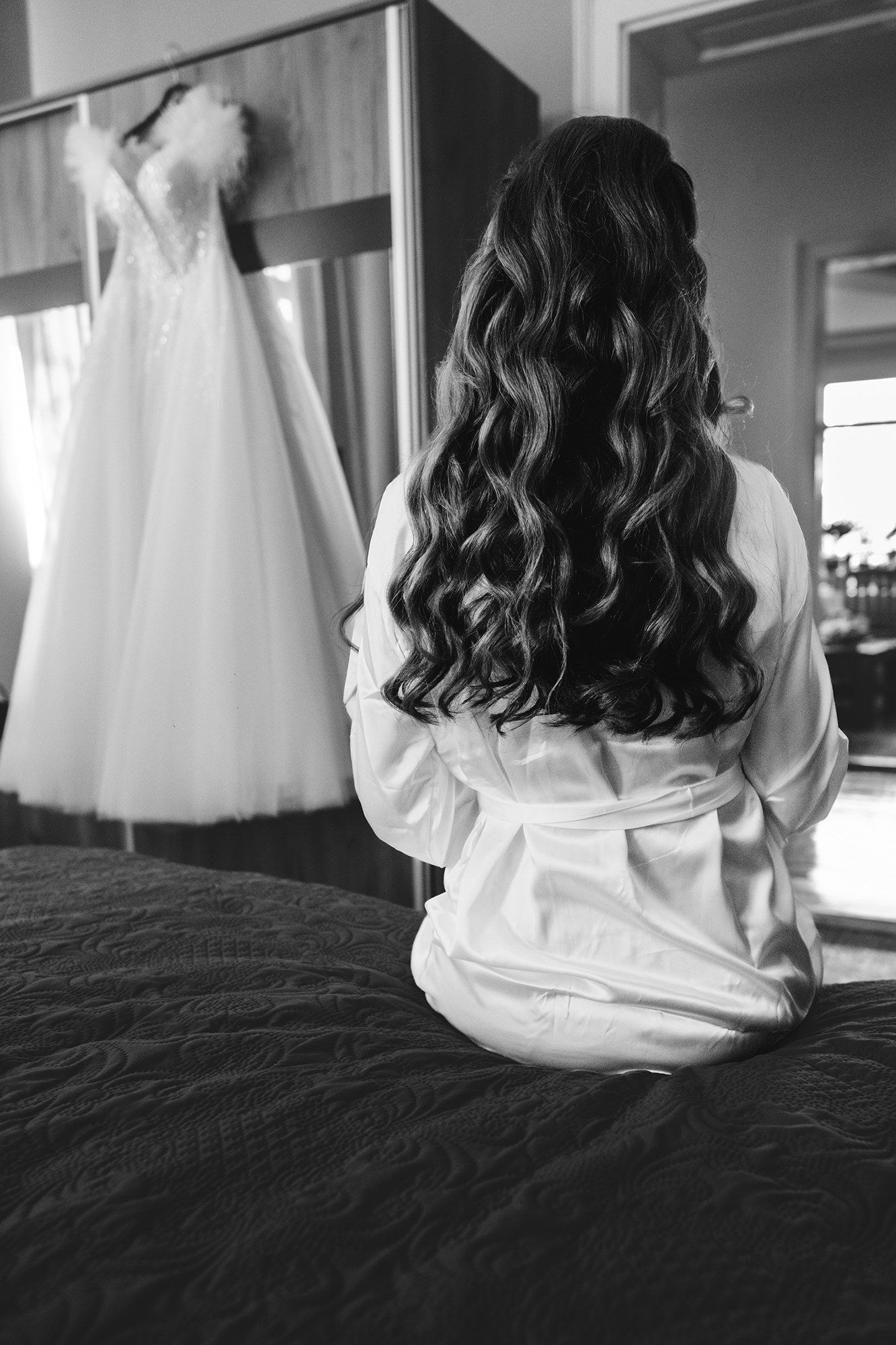 Black and white image of person in white robe sitting on bed during wedding morning preparations.