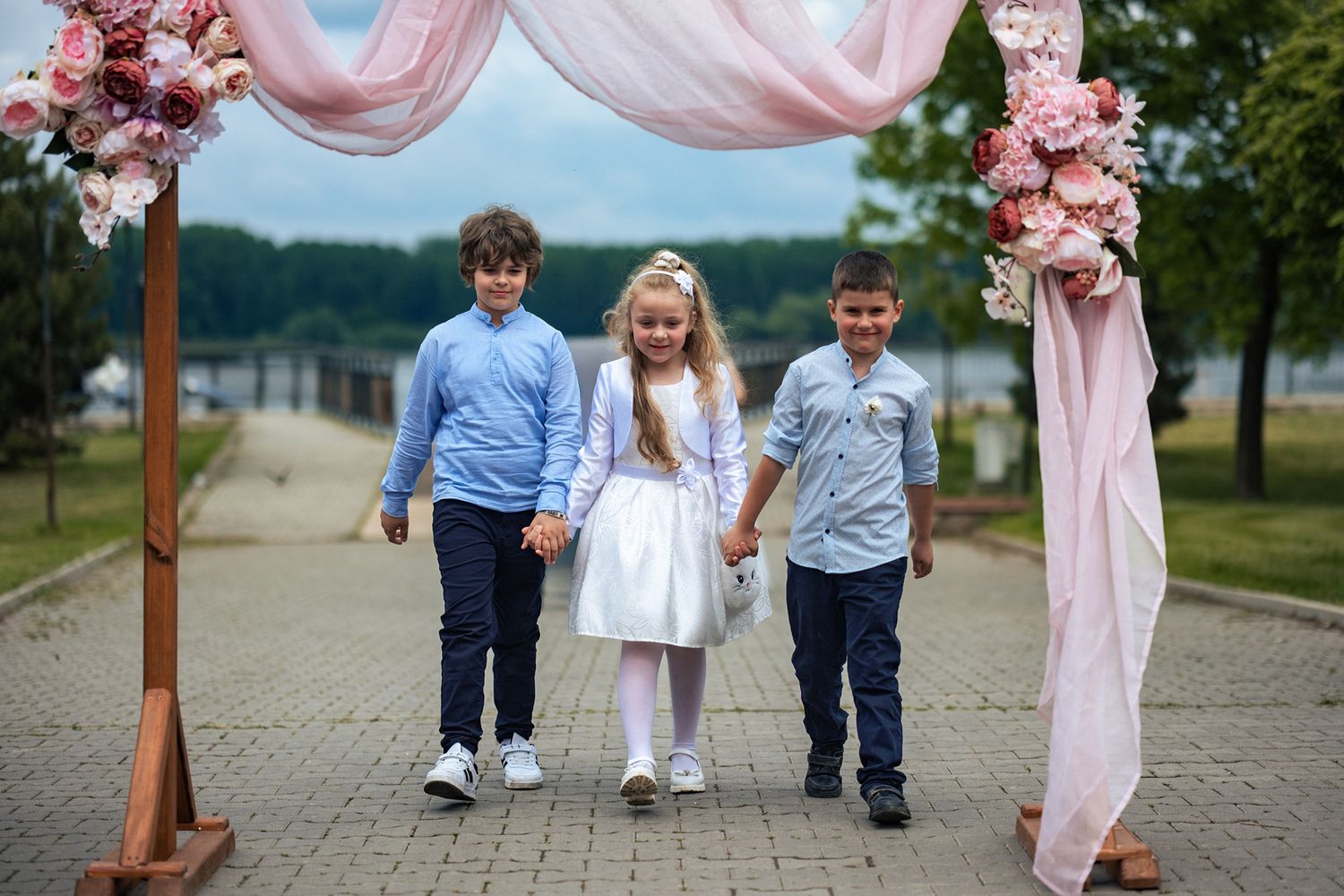 Three children walking under pink draped wedding arch outdoors.