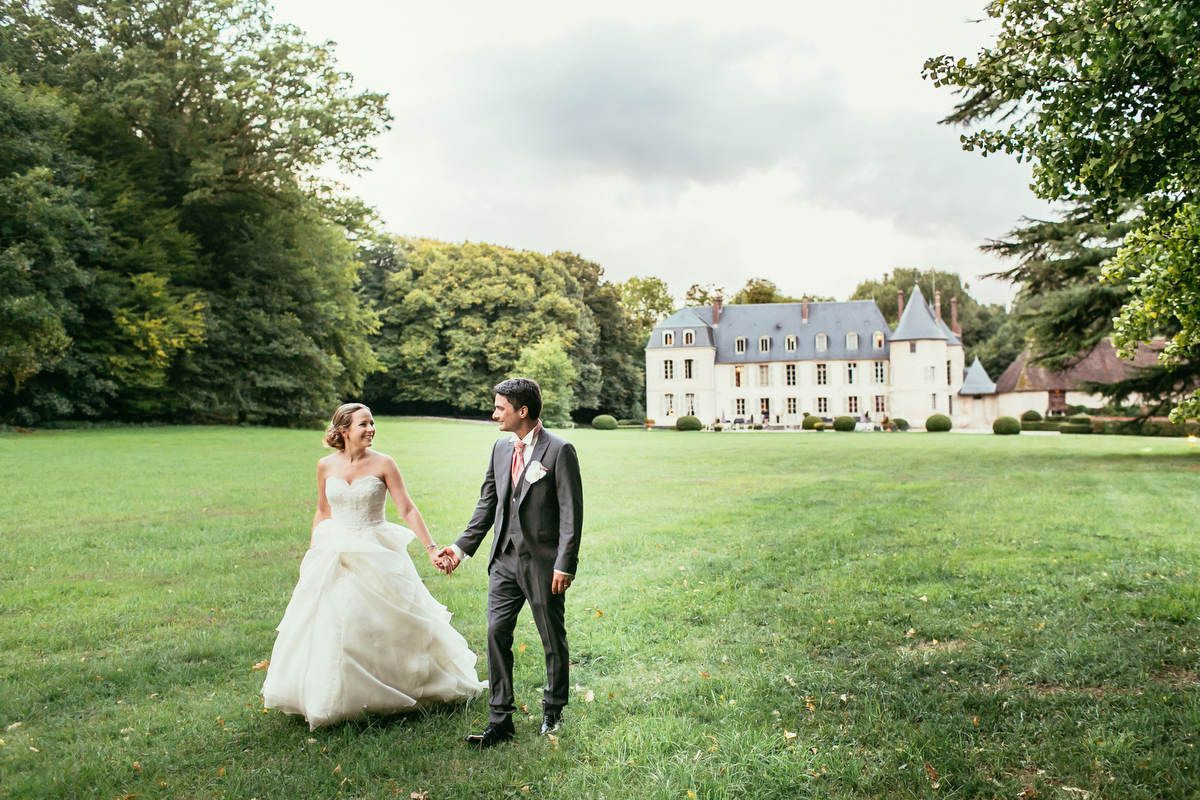Newlyweds walk hand in hand across a sweeping green lawn with the historic Chateau de Changy facade in the background.
