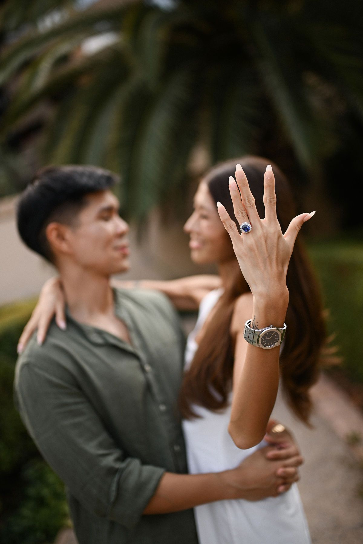 Close-up of a woman's hand displaying a blue engagement ring, with her partner looking at her in the blurred background