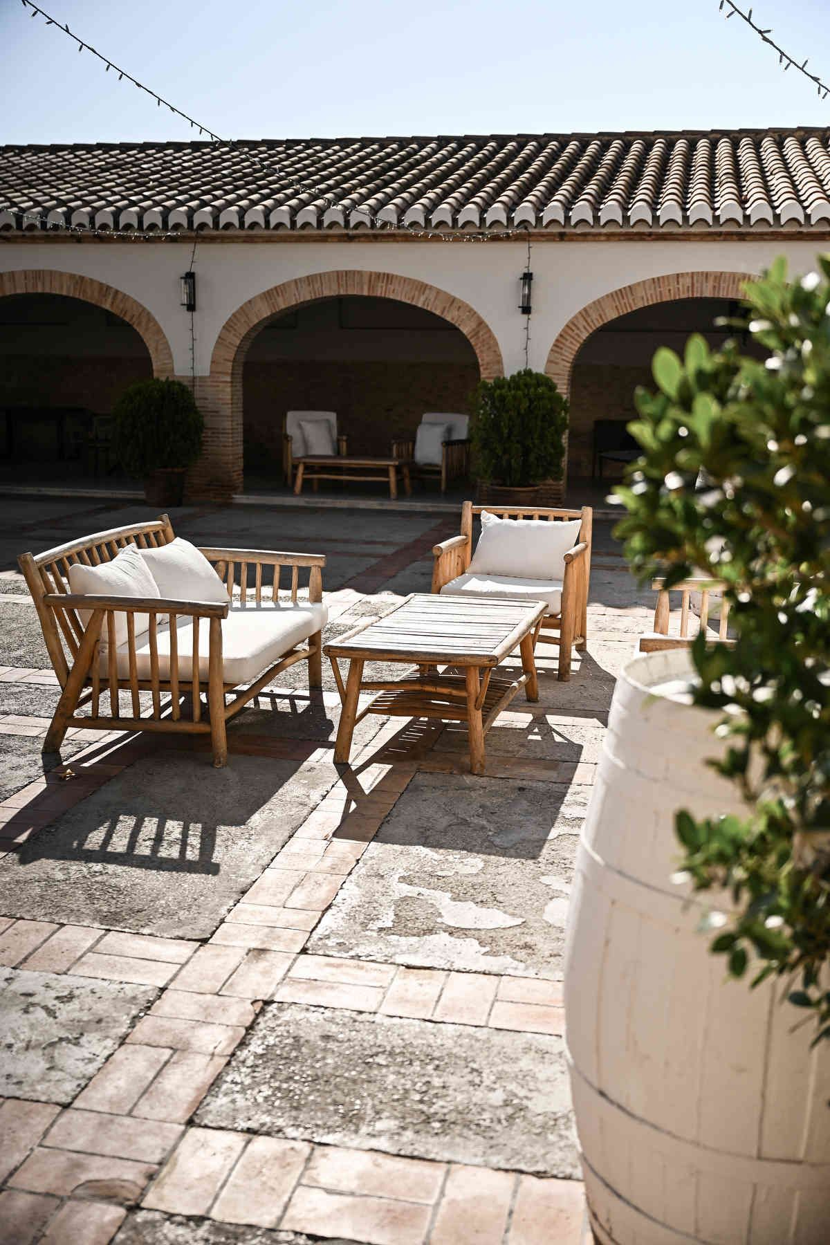 Sunny outdoor patio at Alquería Kukala with wooden sofas, brick arches, and a white decorative barrel.