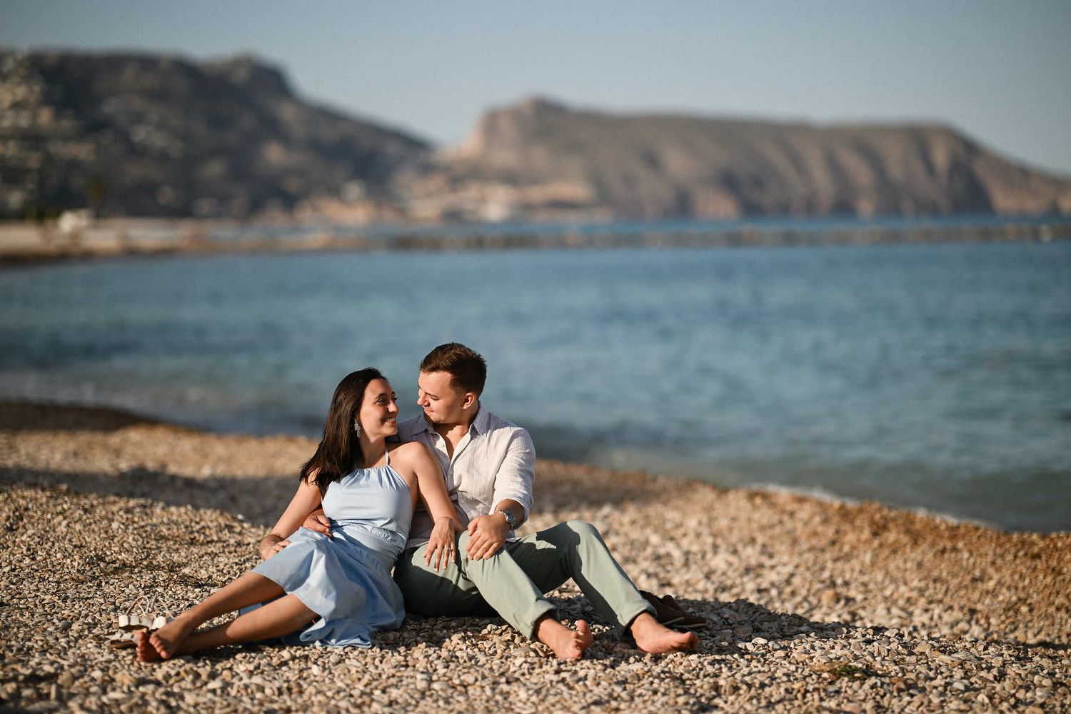A couple sits barefoot on a pebble beach looking at each other near the water following a surprise proposal in Altea.