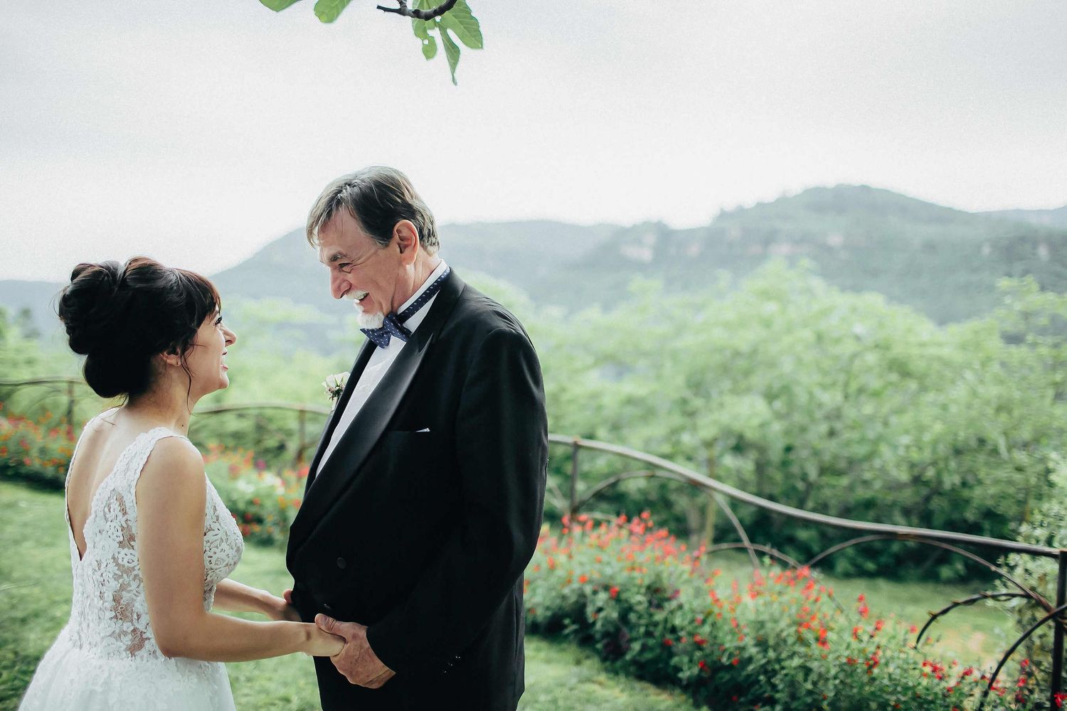A touching moment of a father holding the bride's hands against the scenic mountains at Masia El Follo. Captured by a wedding photographer i