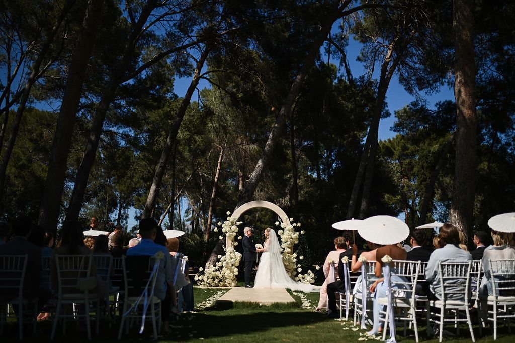 Newlyweds holding hands under a stunning white floral arch surrounded by tall pine trees at a Spanish countryside estate.