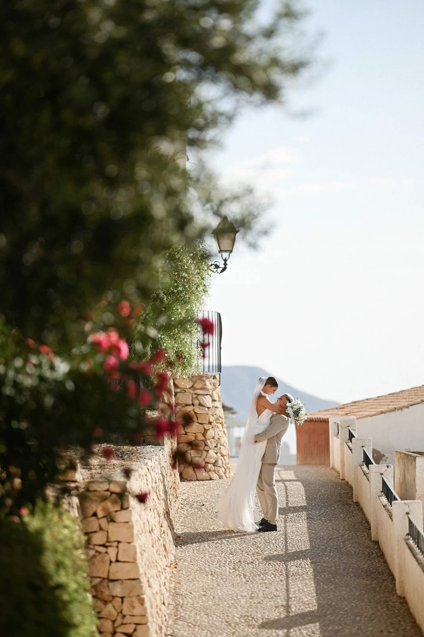 Couple embracing on a stone lane in Altea Old Town with flowers, white walls and the mountains behind.