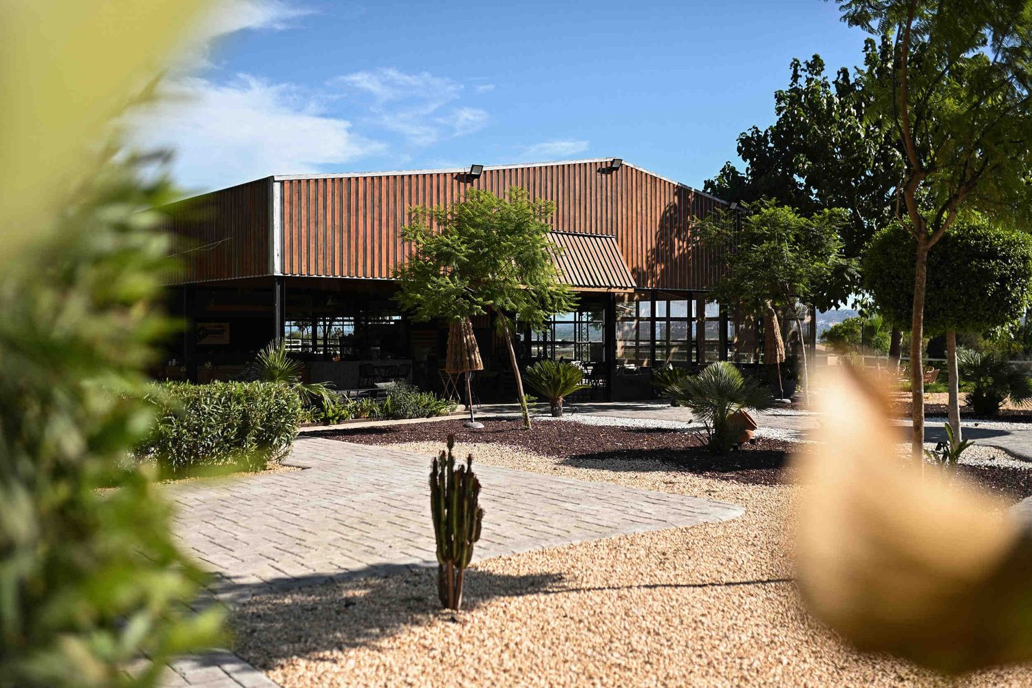 Exterior view of the modern wooden banquet hall at Masia El Graner surrounded by green trees and gravel paths.