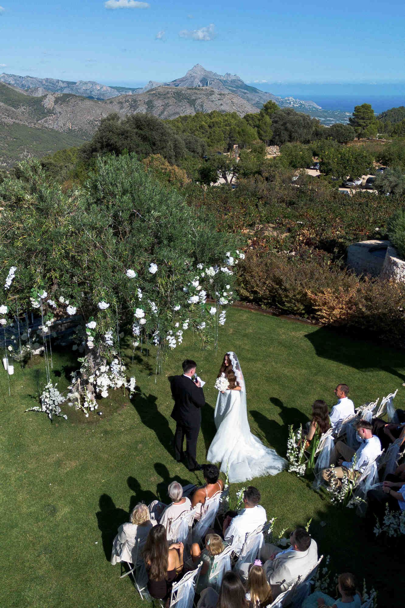 Guests watching the bride and groom exchange vows under a grand floral tree at Masos de Guadalest estate.