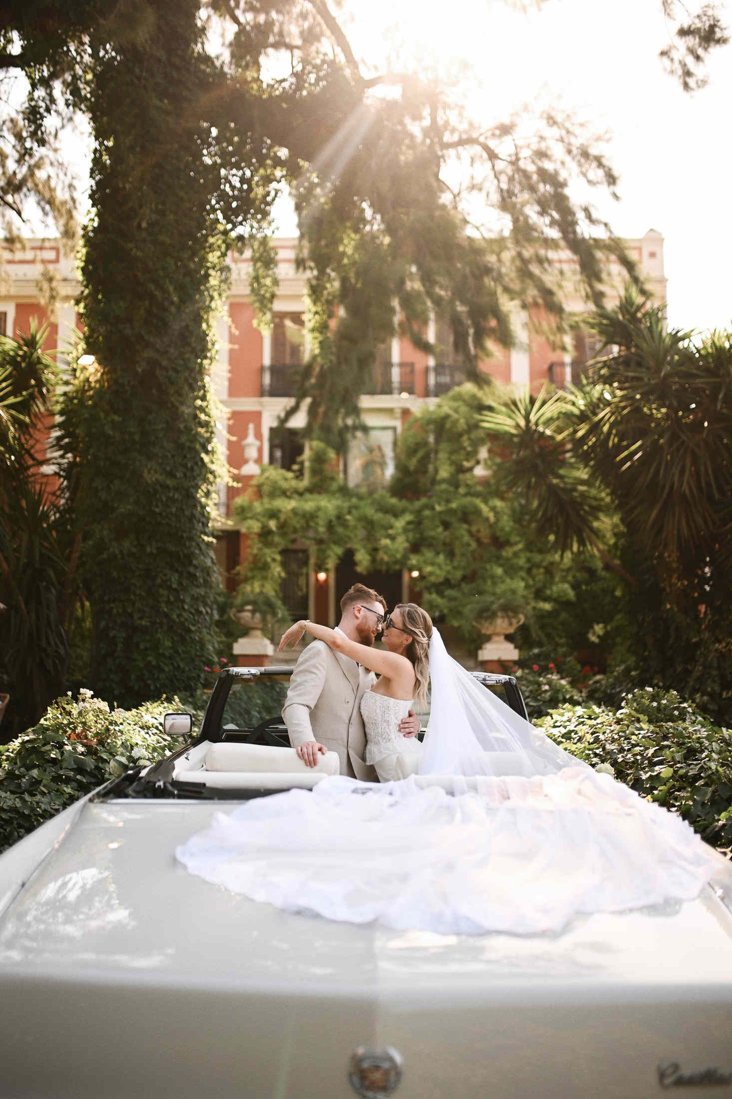 Newlyweds hugging inside a vintage Cadillac with a long wedding veil beautifully spread out by the Villa Dela entrance.