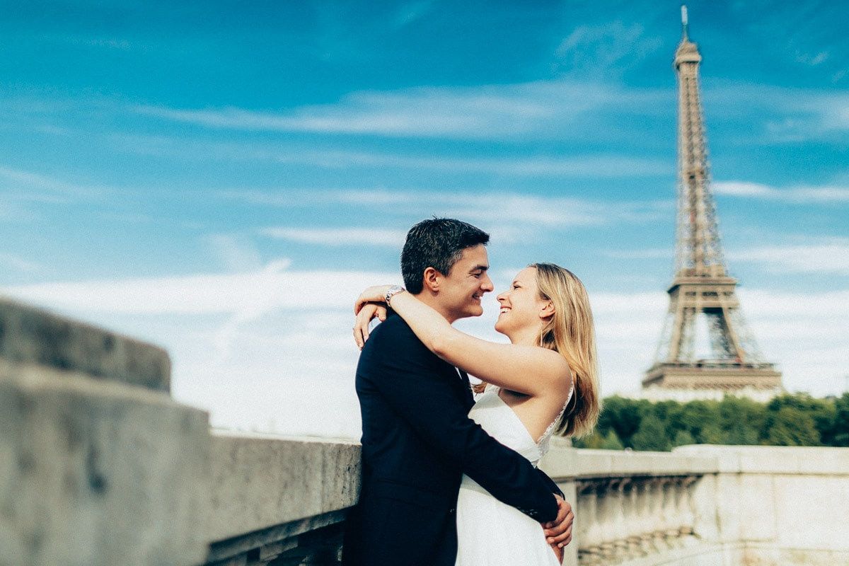 Smiling couple embracing in the sunlight with the Eiffel Tower and a clear blue sky behind them during a Paris photoshoot.