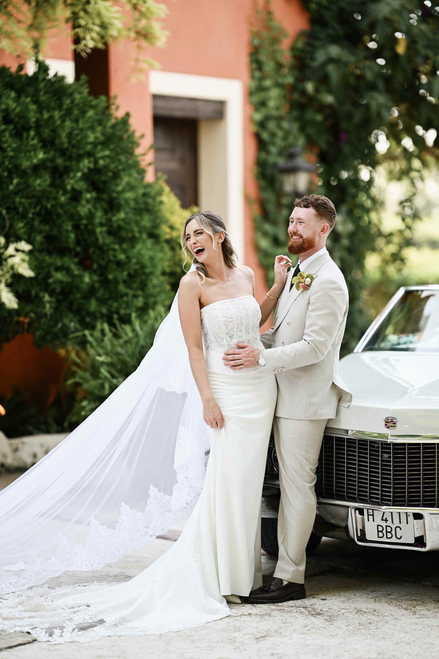 Newlyweds with a retro Cadillac near the terracotta brick facade of Villa Dela destination wedding venue in Spain.