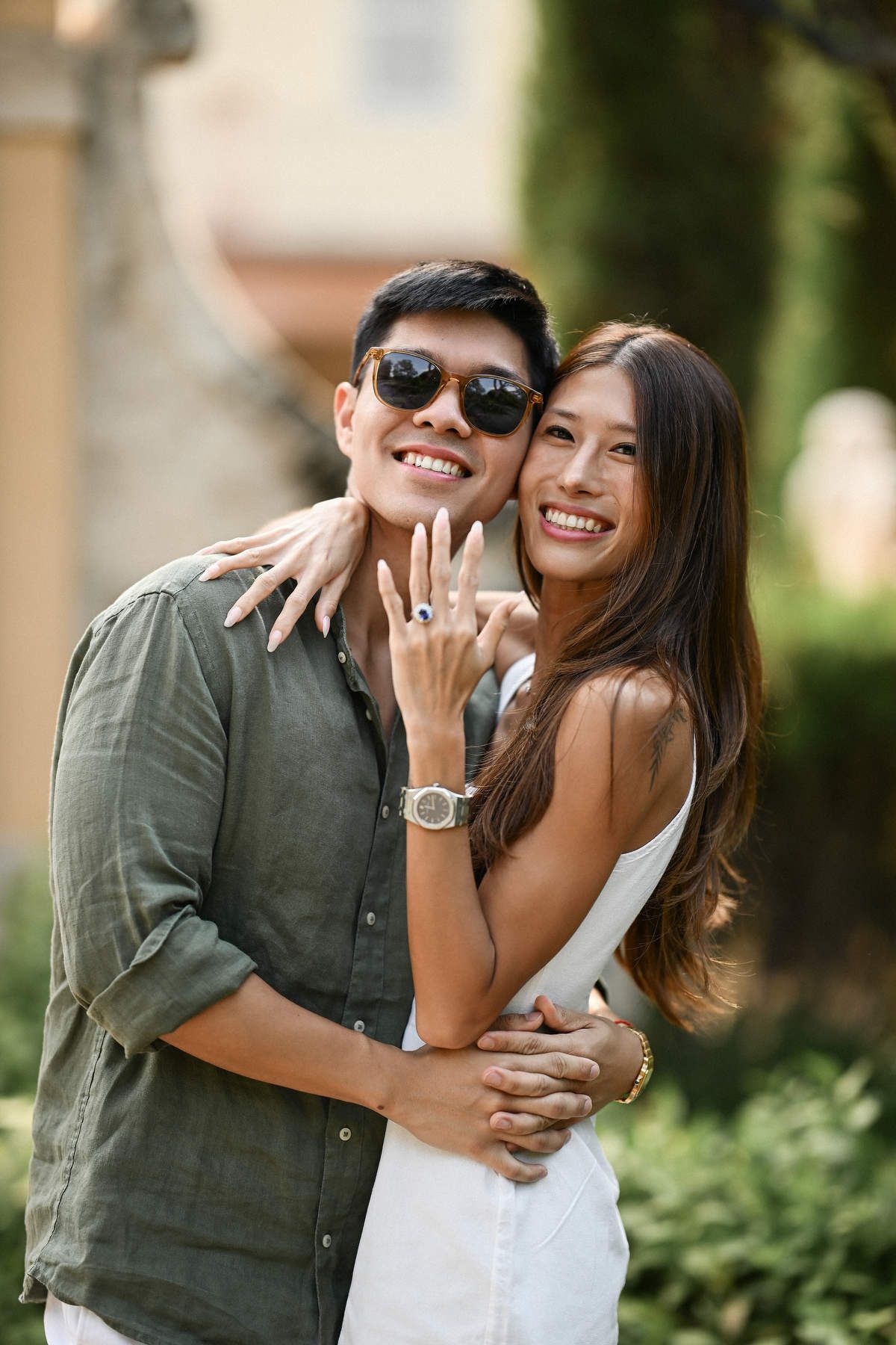 A smiling woman shows off her new blue gemstone engagement ring while hugging her partner outdoors.