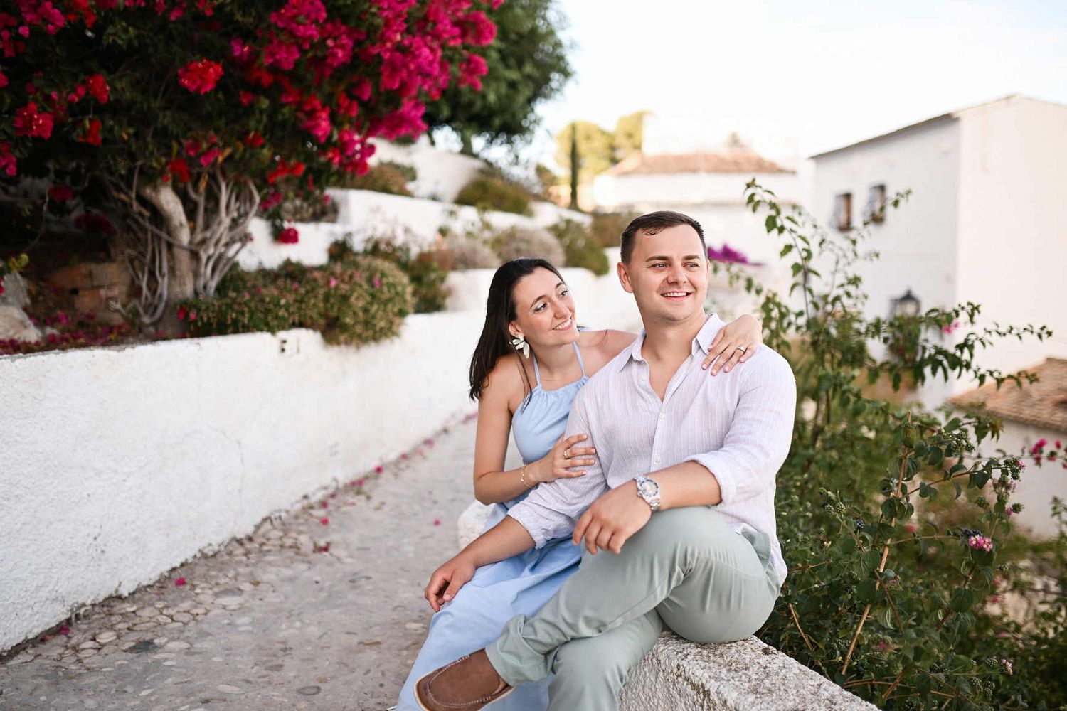 A man and woman sit together on a low stone wall near pink bougainvillea flowers during their Altea proposal photoshoot.