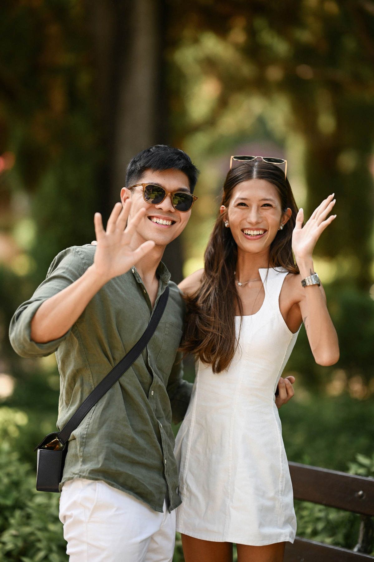 Smiling couple waving at the camera after a secret proposal in the shaded green areas of Monforte Gardens.