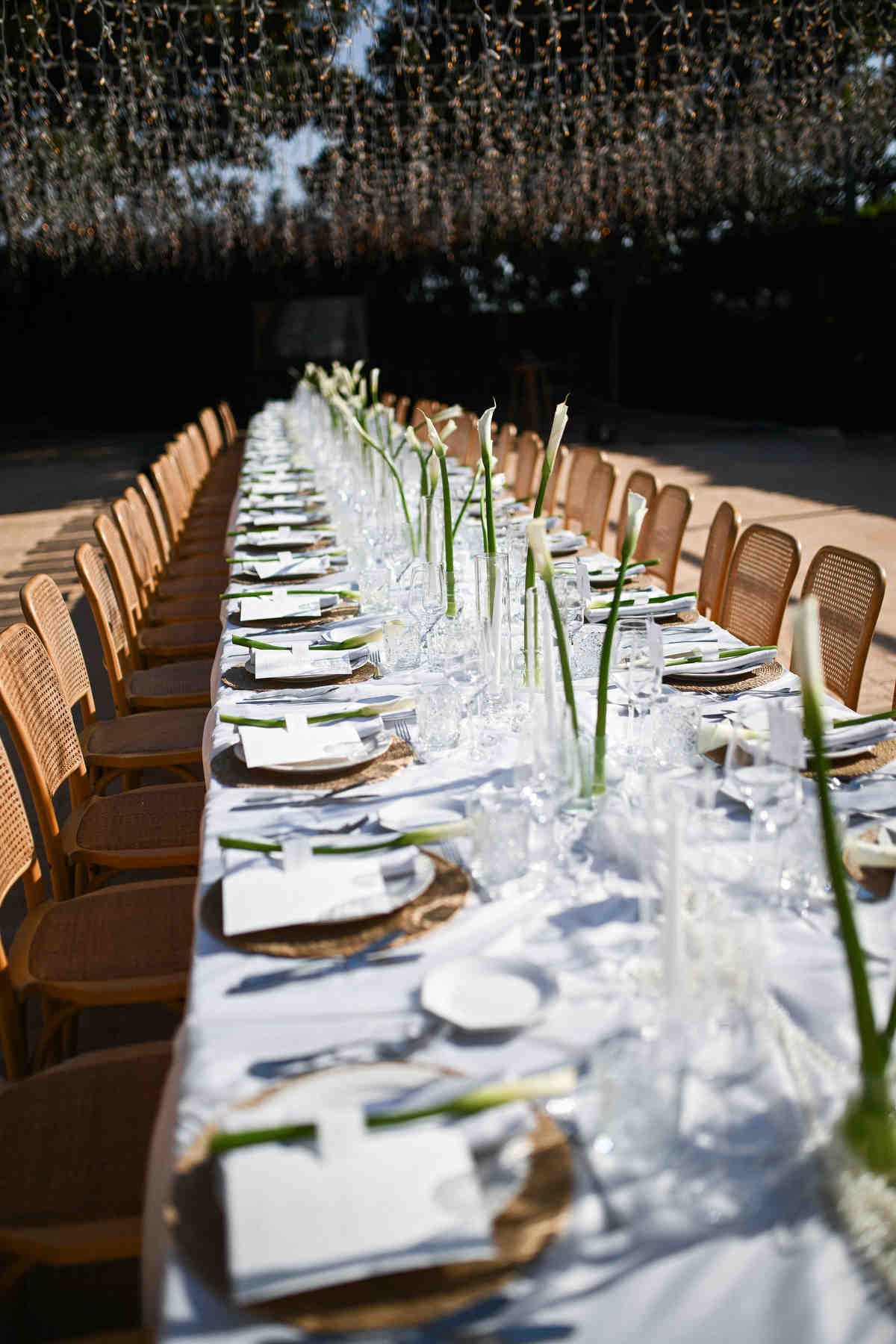 Long outdoor wedding banquet table with white tablecloth, wooden chairs, and calla lilies under string lights at Alquería Kukala.