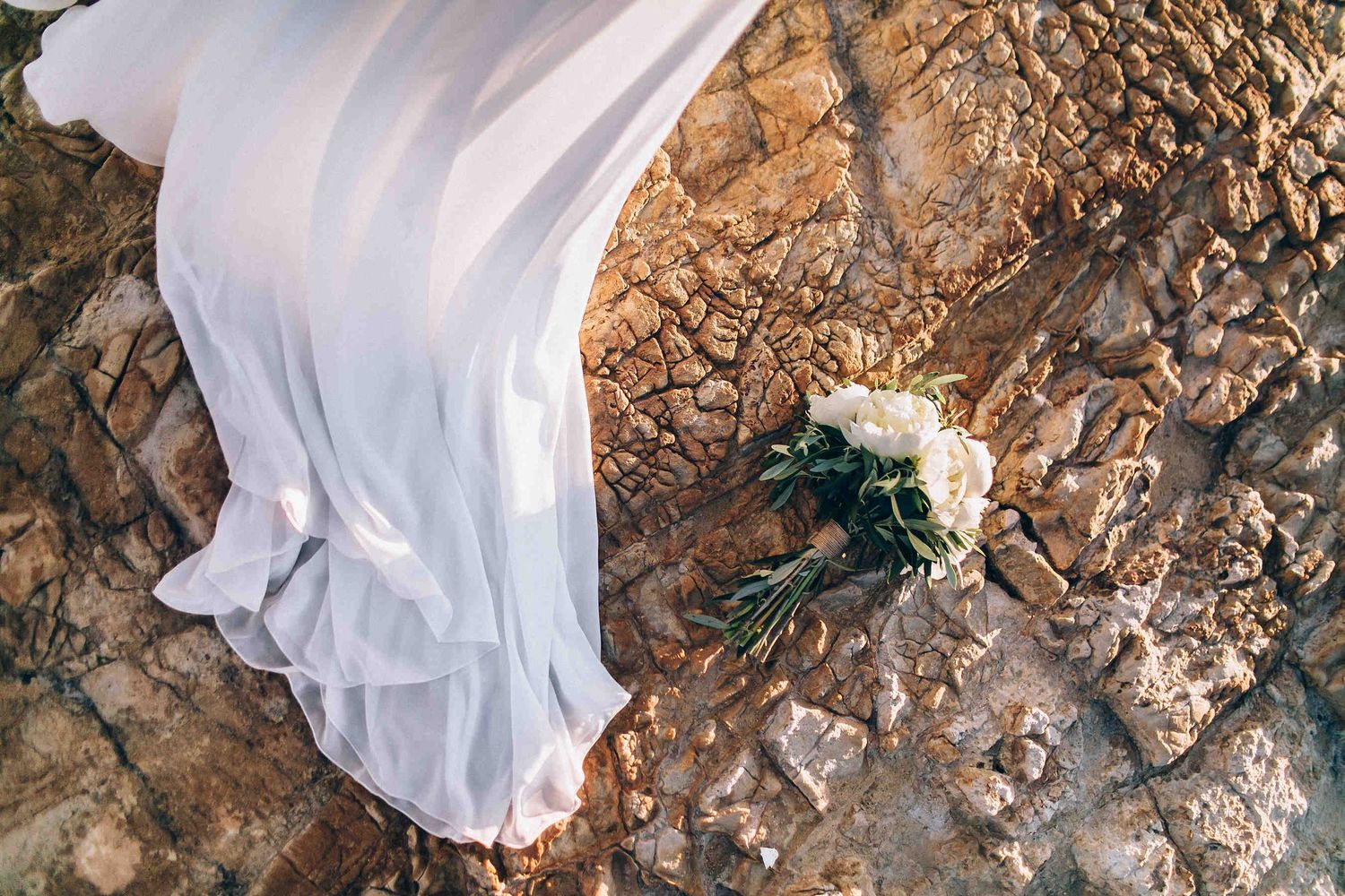 Creative shot of the bridal bouquet resting on the raw volcanic rock and silk wedding dress