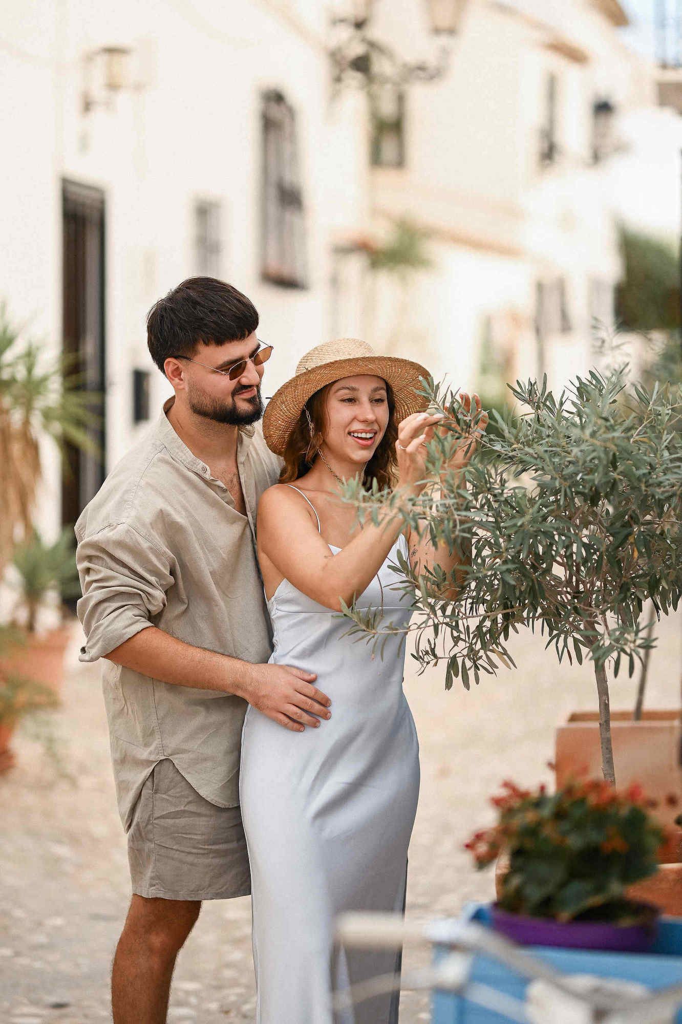Pre-wedding photo session Spain. A man embraces a woman as she touches a fresh olive on a tree in a picturesque Spanish location.