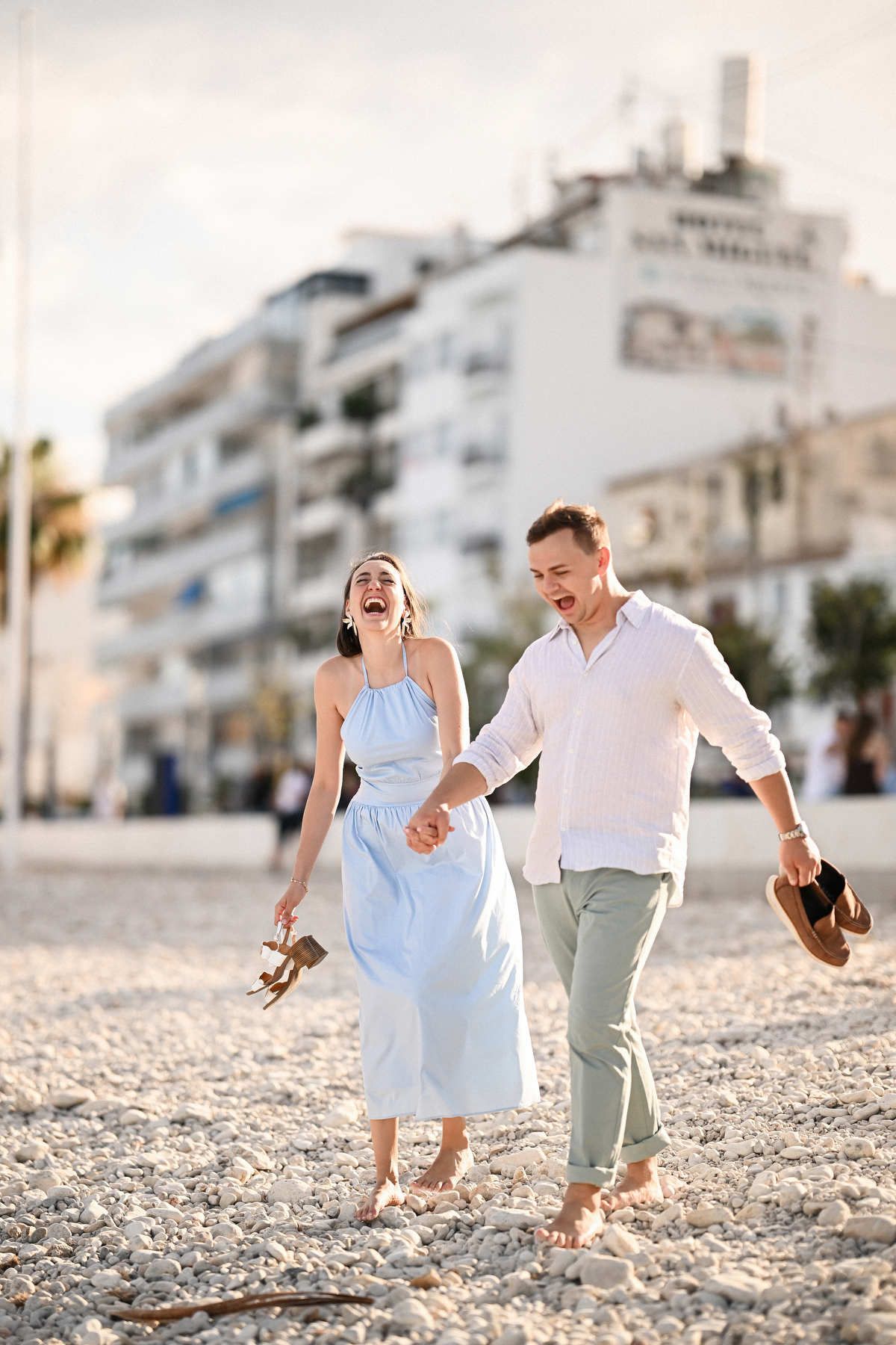 An Altea proposal photographer captures a barefoot couple holding their shoes and laughing on a bright pebble beach.