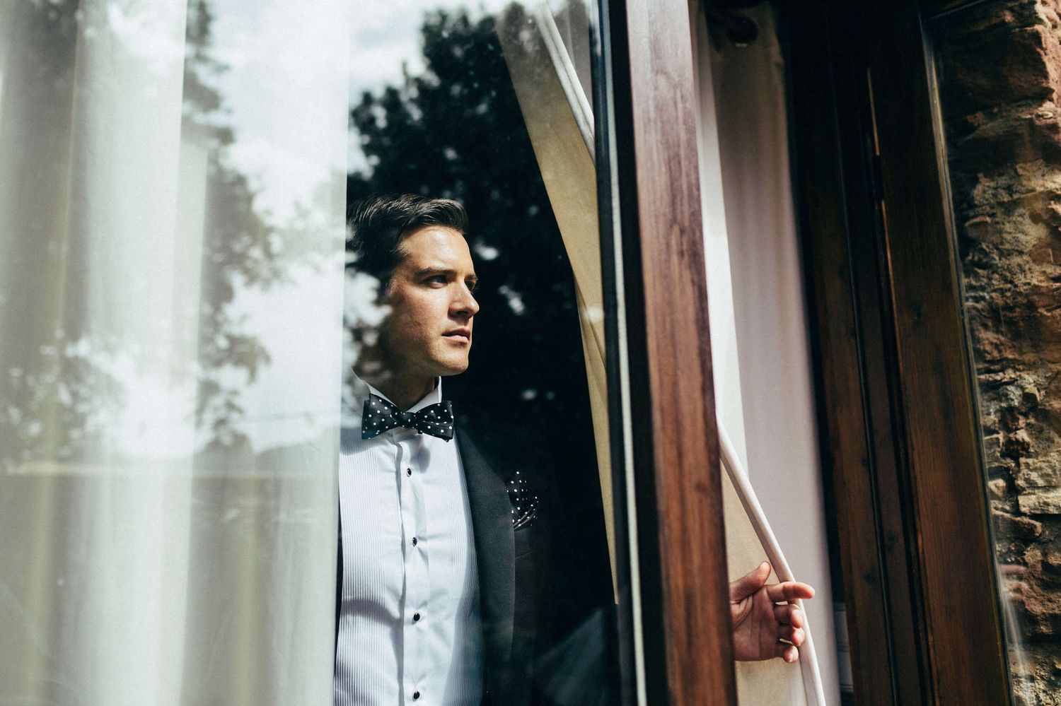 Cinematic shot of the groom looking through a European villa window, watching the bride's arrival for an intimate wedding ceremony.