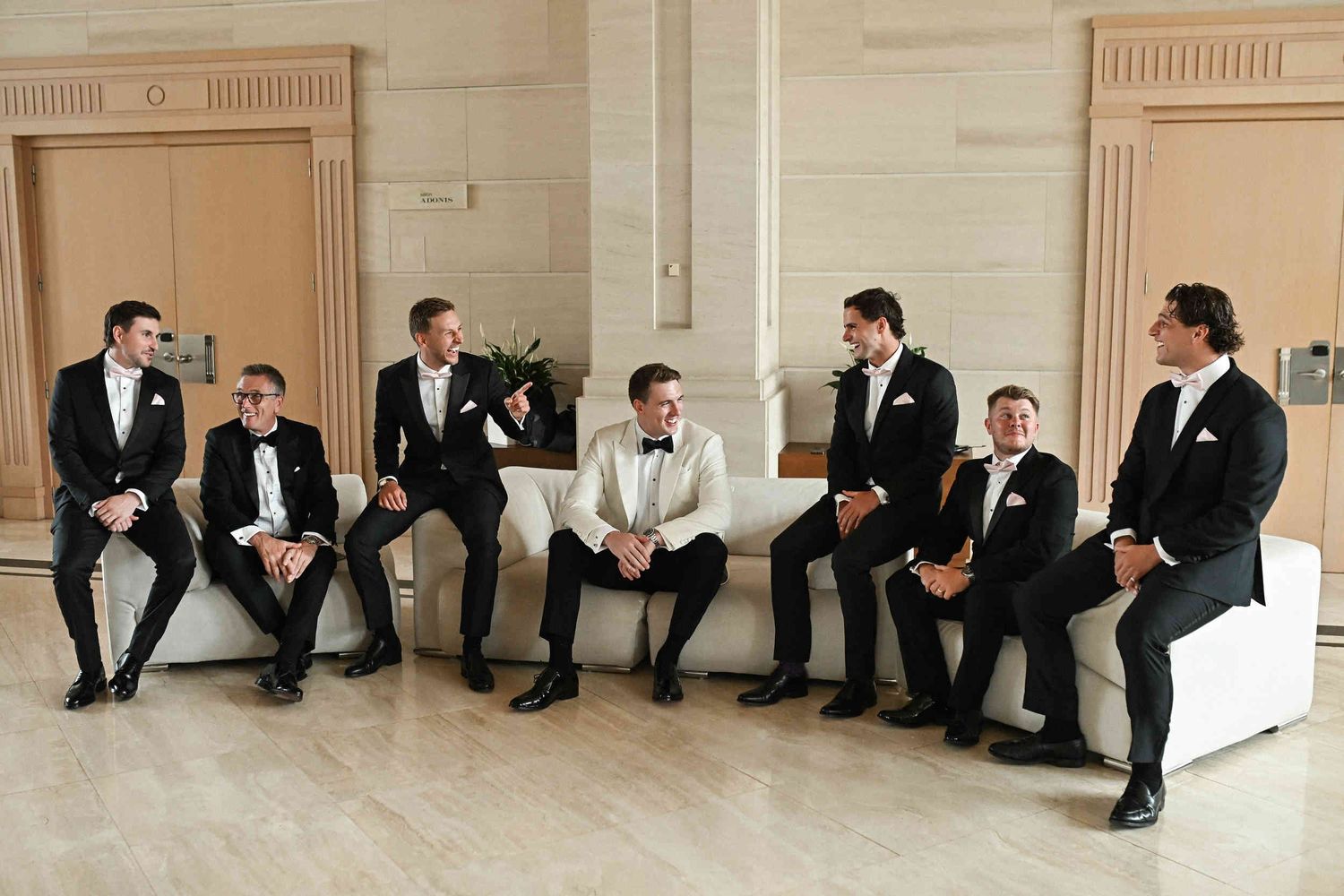 Groom in a white tuxedo laughing with groomsmen seated on white sofas in a spacious hotel lobby.