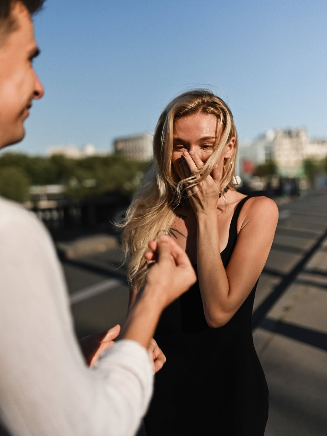 Joyful woman covering her mouth in shock as her partner presents an engagement ring in bright evening sunlight in Paris.