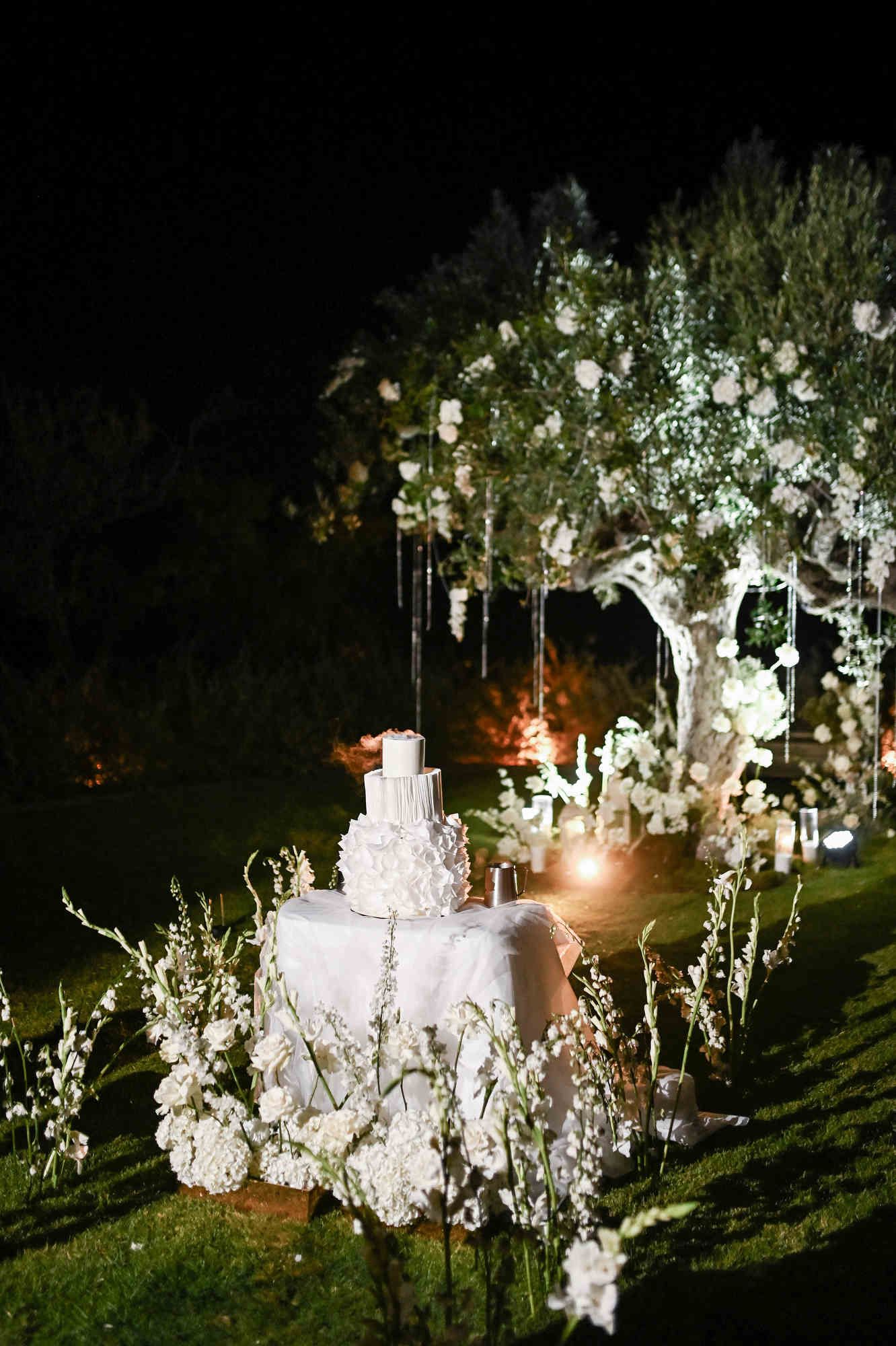White tiered wedding cake on a illuminated table under a decorated olive tree at night in Bodega MASOS.