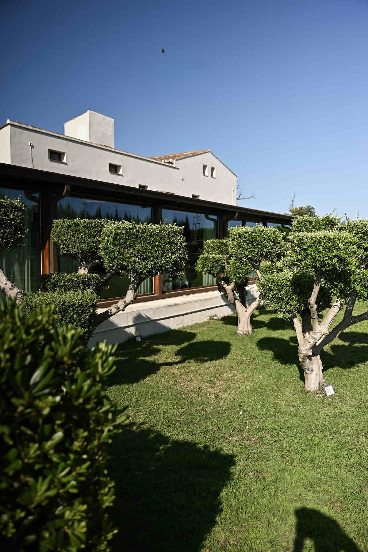 Main salon building at Alquería Kukala with large panoramic windows and neatly shaped olive trees on green grass.