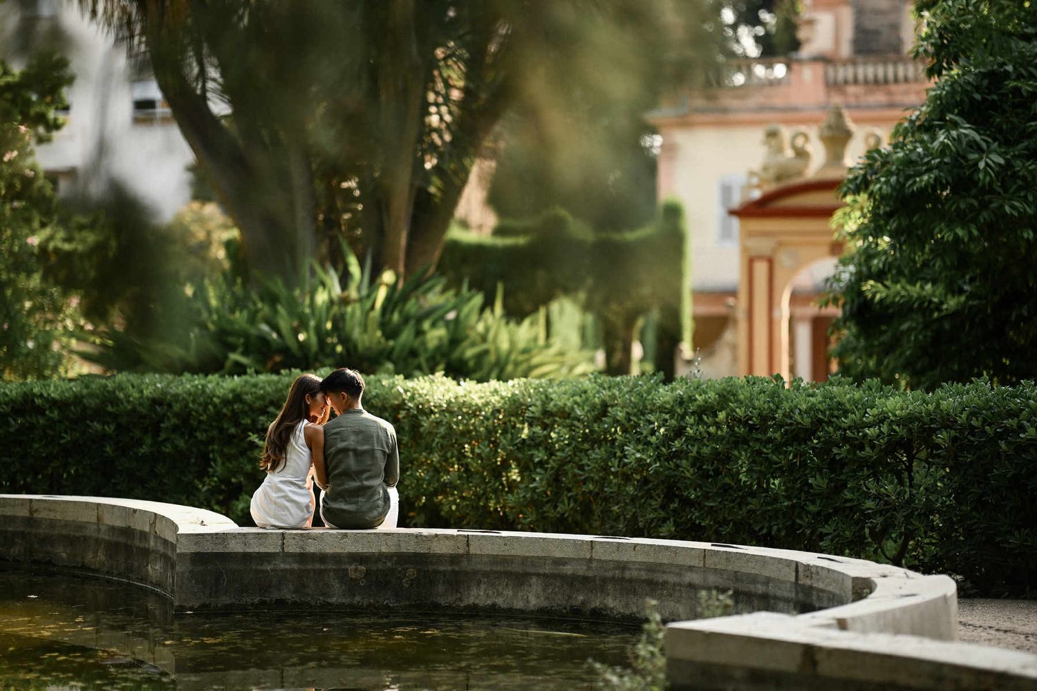 A couple sits closely on a curved stone wall beside a calm pond after deciding to propose in Valencia gardens.