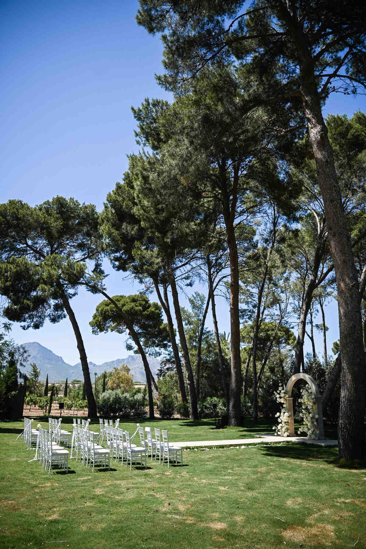 Outdoor wedding ceremony setup on a green lawn with white chairs and a floral arch placed directly under large pine trees.