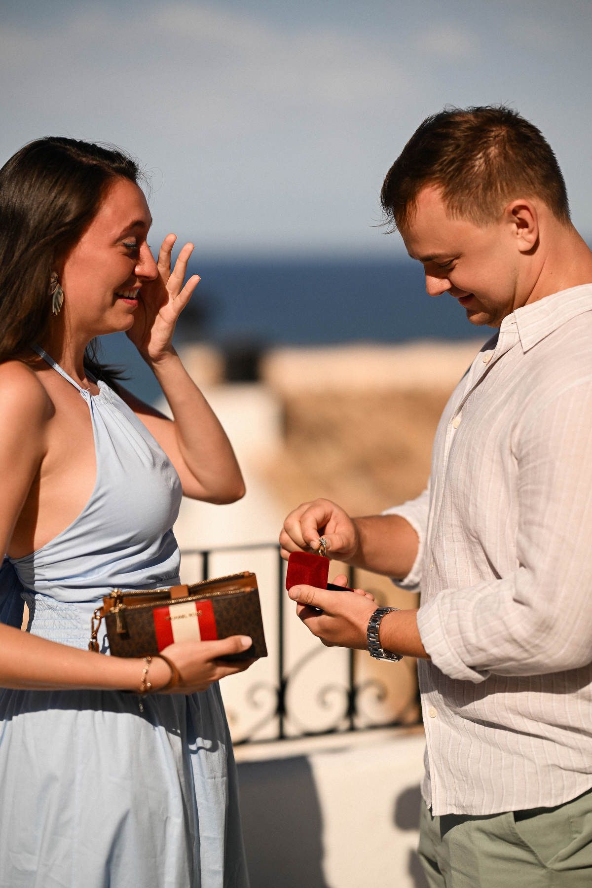 A woman wipes a tear from her eye as her partner holds a ring box during their secret proposal by the sea in Altea.