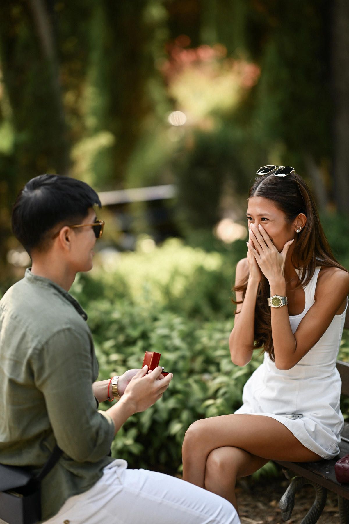 A surprised woman covers her mouth in pure shock as her partner opens a red ring box on a neoclassical park bench.