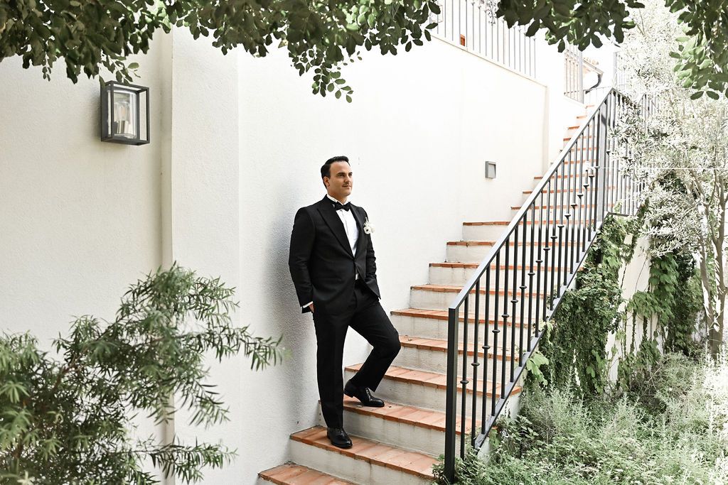 Full-length cinematic portrait of the groom in a tailored black suit posing on the historic stone staircase at Finca del Canónigo.
