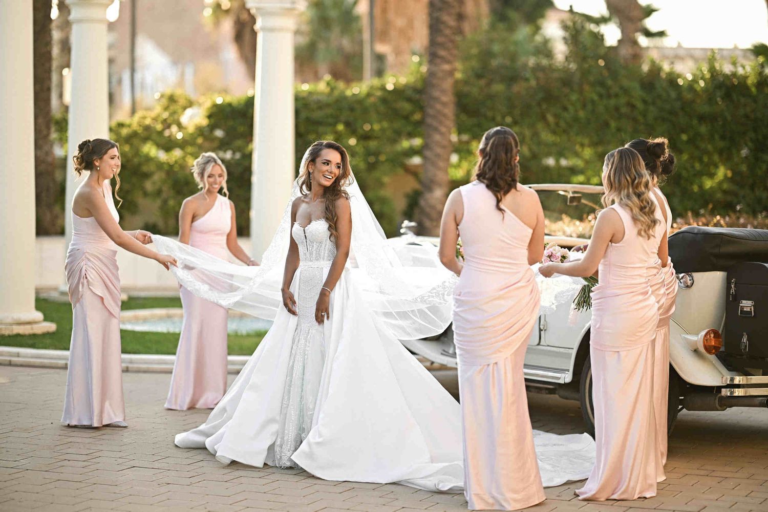 Bridesmaids helping a bride in a mermaid gown with her long veil next to a vintage car in Hotel Las Arenas.